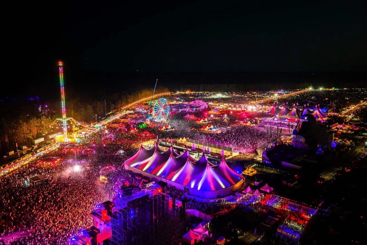 The site at night from above in 2026: lit-up big tops, Ferris wheel, drop tower and crowd in front of the Mainstage.