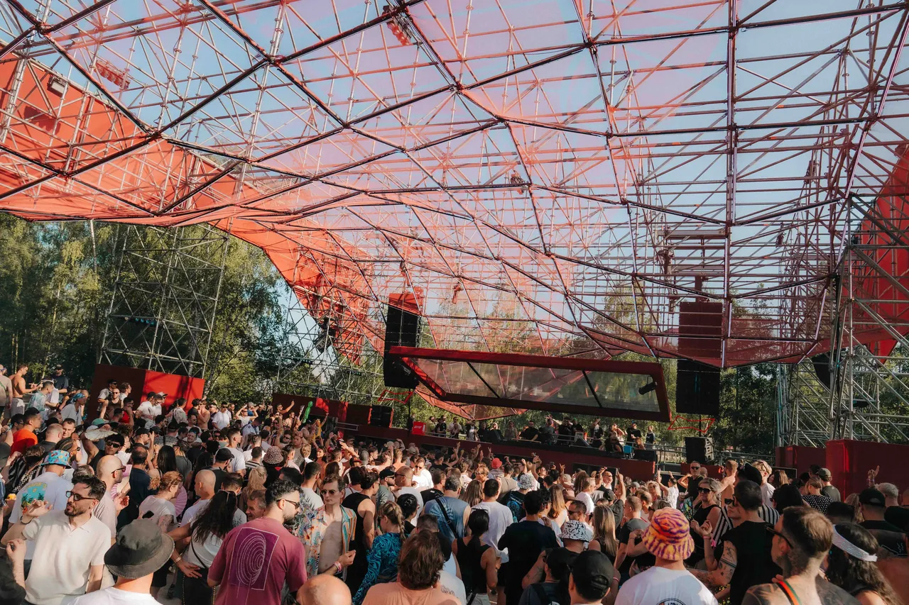 Une area sous une charpente métallique tendue de toile rouge, en plein jour, en juillet 2026.