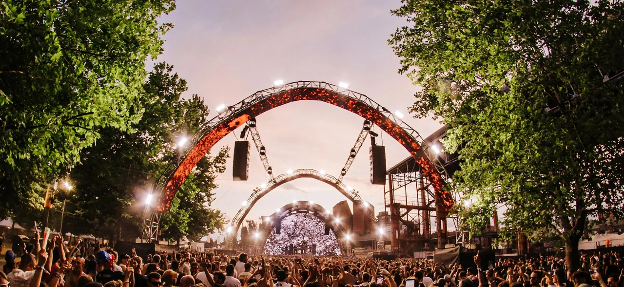 A stage framed by illuminated arches, between the park's trees, at dusk.