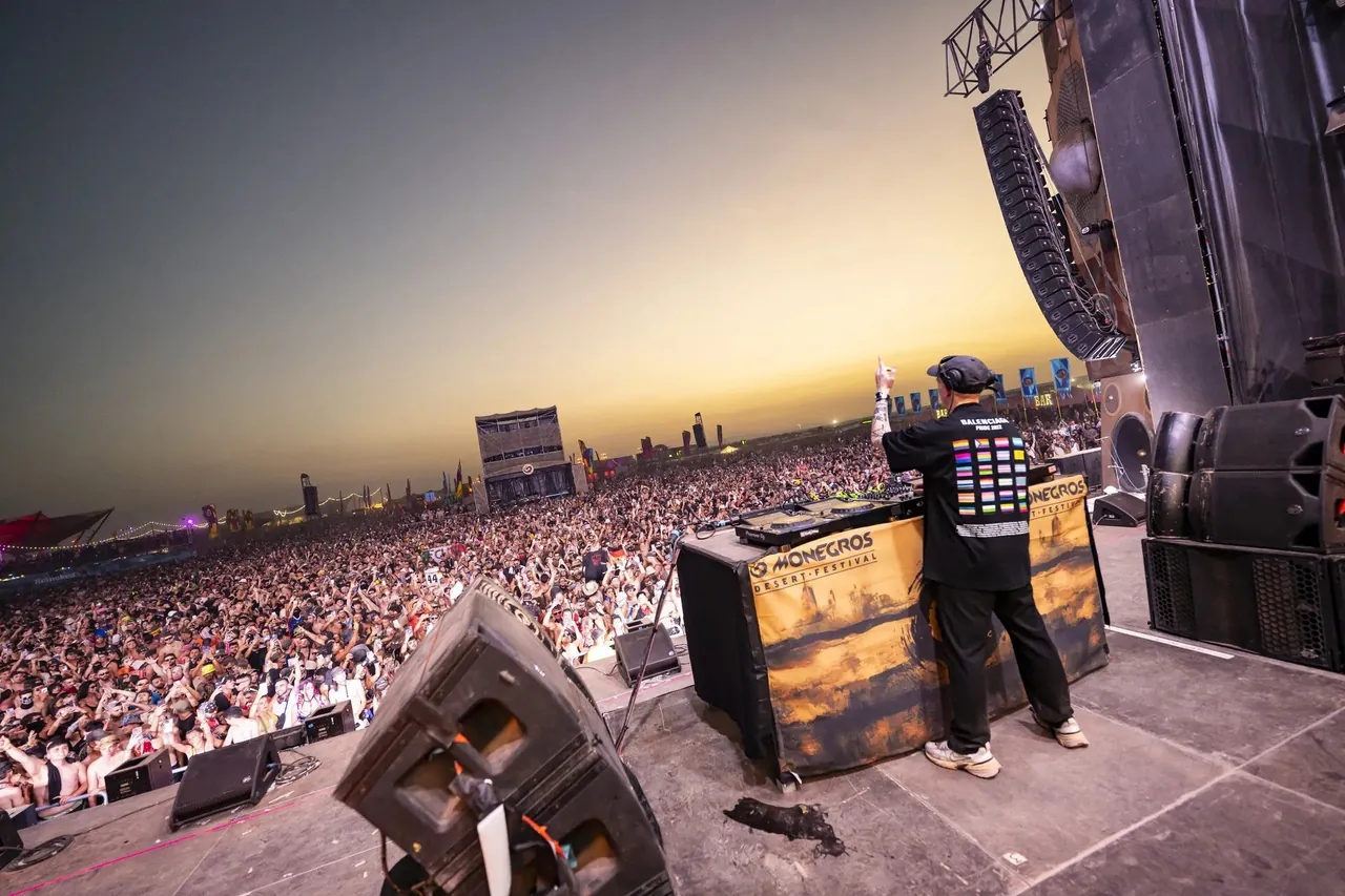 A DJ facing the crowd at sunset, seen from behind the stage.