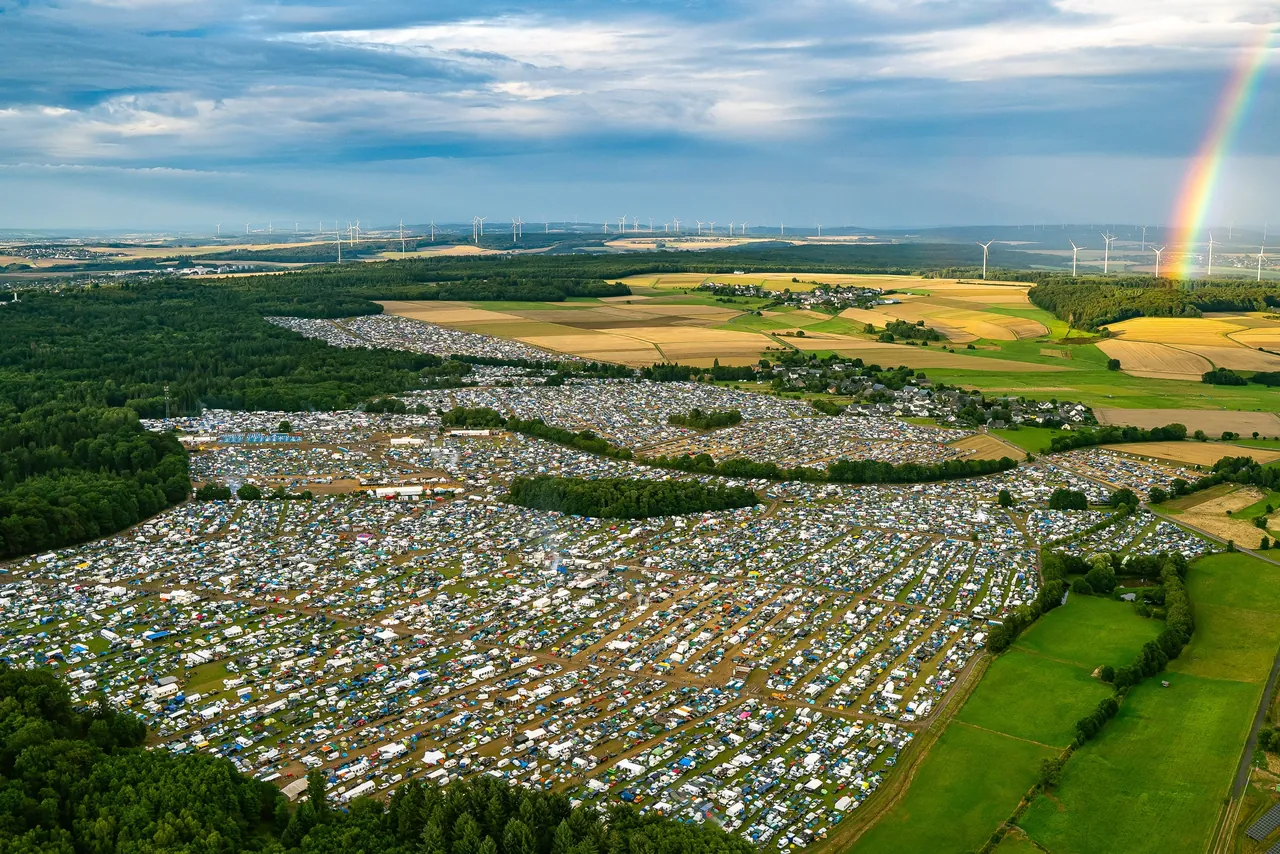Le CampingVillage vu du ciel en 2023 : des milliers de tentes et de voitures entre forêt et champs du Hunsrück, sous un arc-en-ciel.