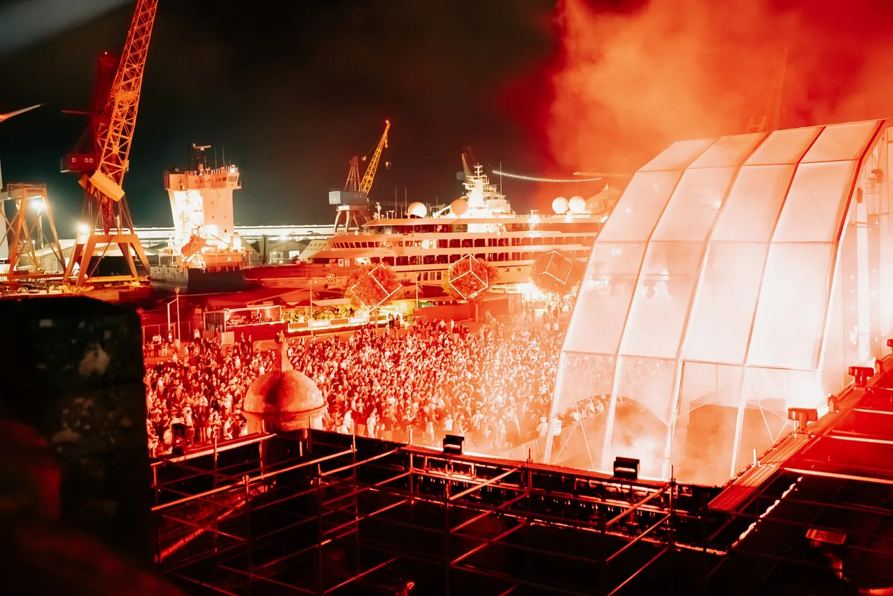 The Neo Stage crowd in red smoke on the night of 6 to 7 August 2026, with cranes and a docked cruise ship behind.