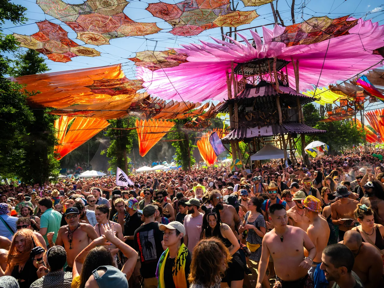 The Ozora Stage in broad daylight: its wooden tower under a canopy of pink and orange fabric, above a dense crowd, in 2023.