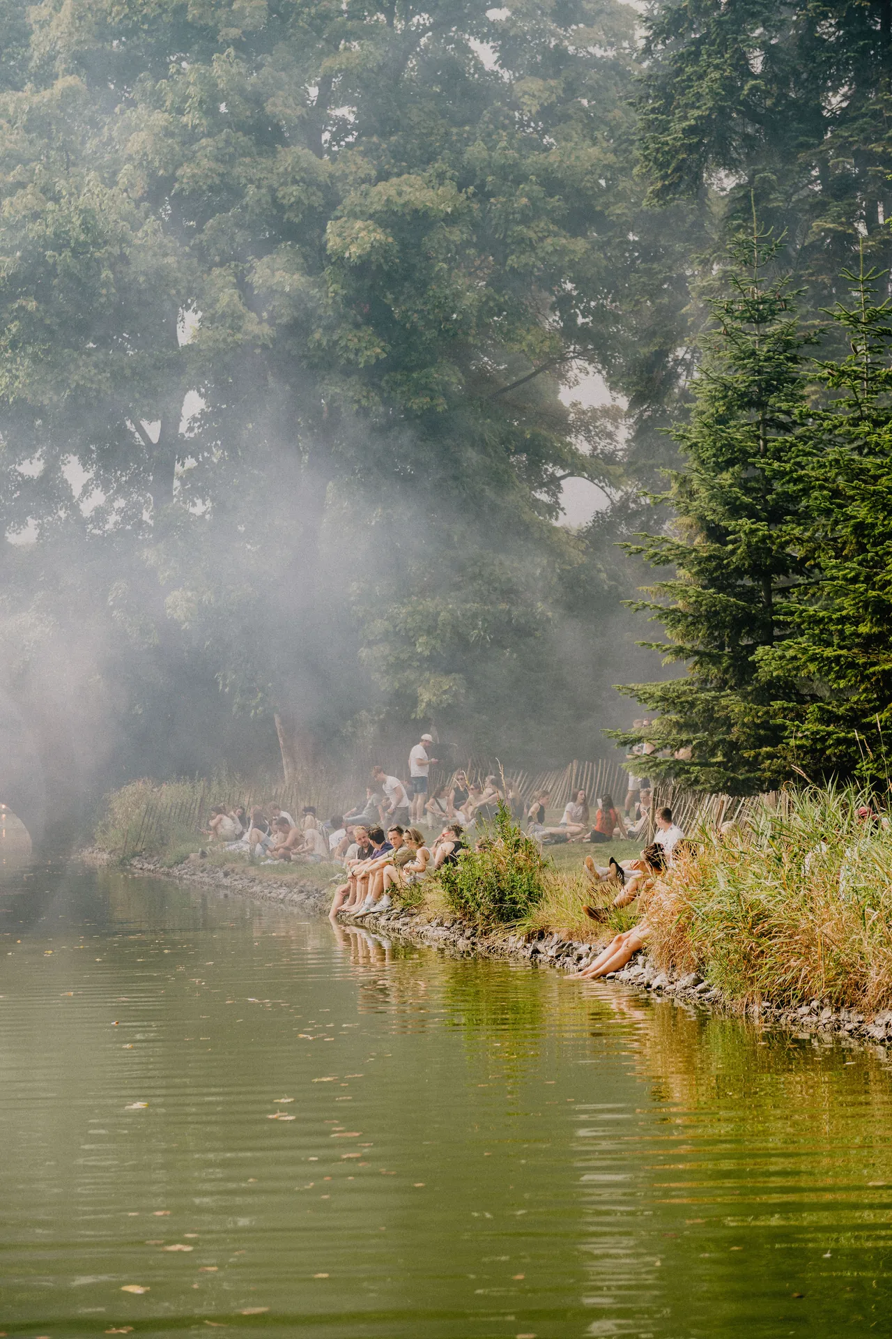 Festivaliers sur la rive du lac, dans la brume et la lumière filtrée par les arbres.