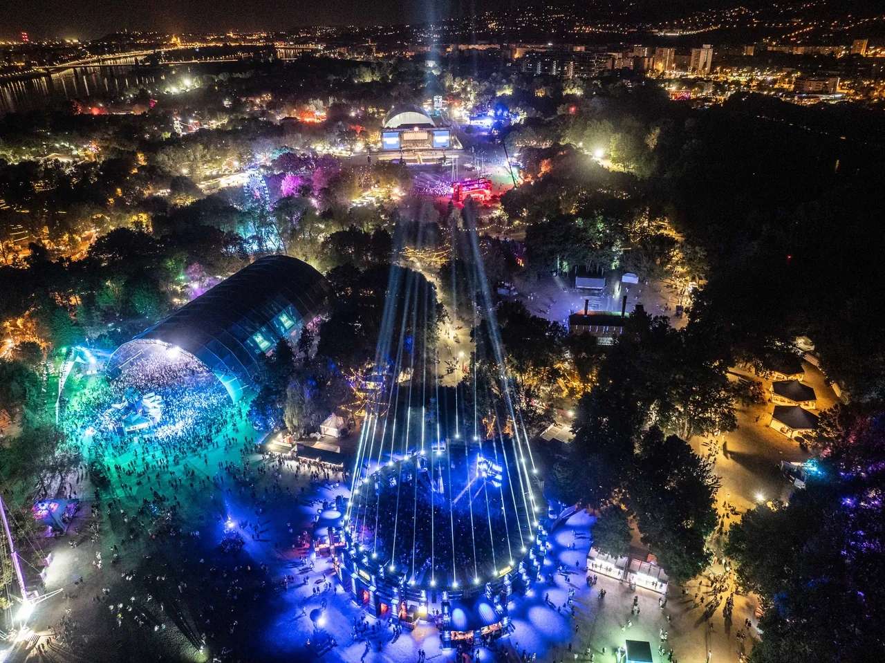 L'île de nuit vue du ciel, le 15 août 2026 vers 1 h : la grande structure couverte de la Revolut Stage à gauche, l'anneau lumineux du Yettel Colosseum au premier plan, le Danube et Budapest au fond.