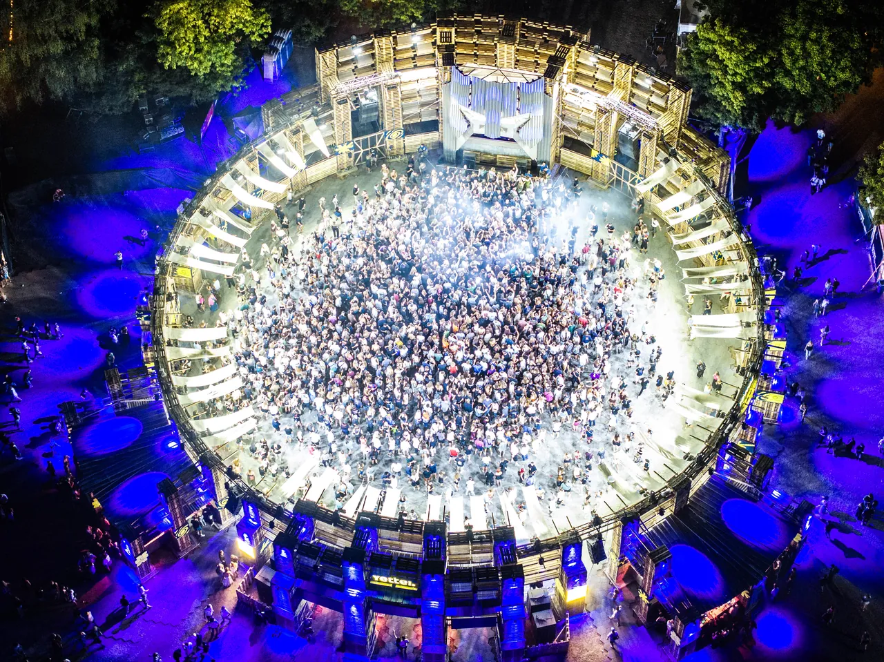 Le Yettel Colosseum vu du ciel dans la nuit du 14 au 15 août 2026 : l'arène en palettes et sa piste de danse circulaire, la cabine du DJ en haut de l'image.