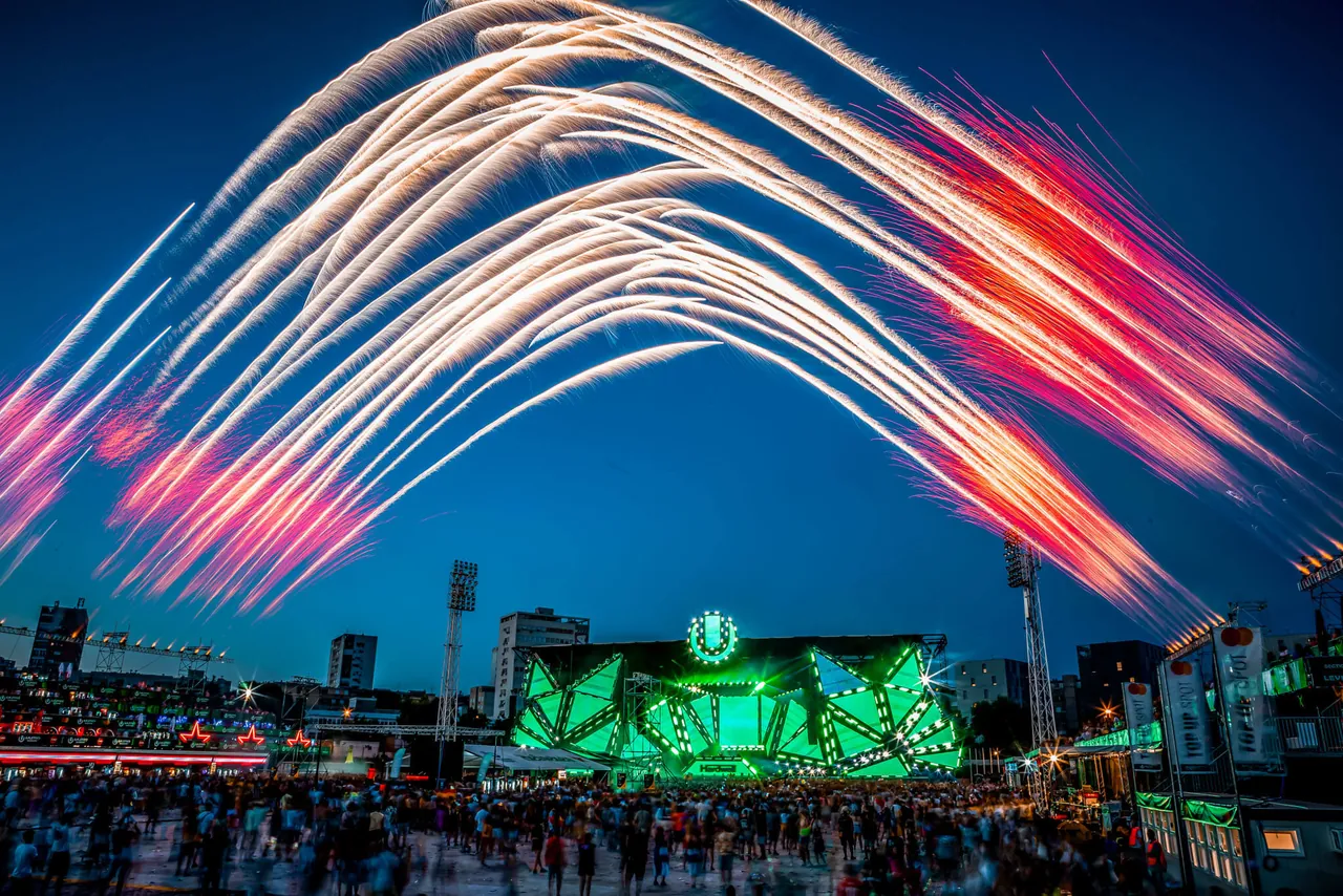 Gerbes de feu d'artifice en arc au-dessus du Mainstage éclairé en vert, à l'heure bleue, en 2023.