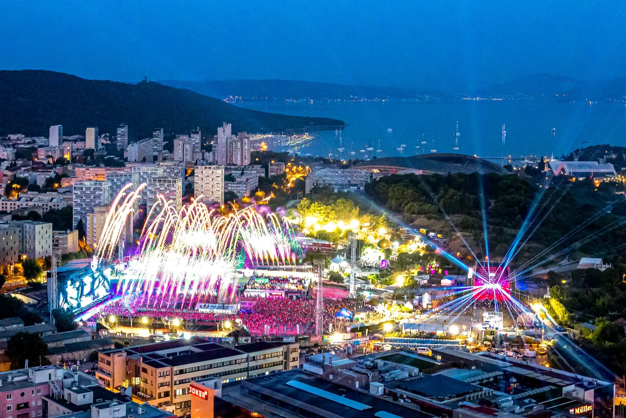 Park Mladeži vu d'en haut à la tombée de la nuit pendant l'édition 2024 : feu d'artifice sur le Mainstage, lasers de la scène voisine, et la baie de Split en arrière-plan.