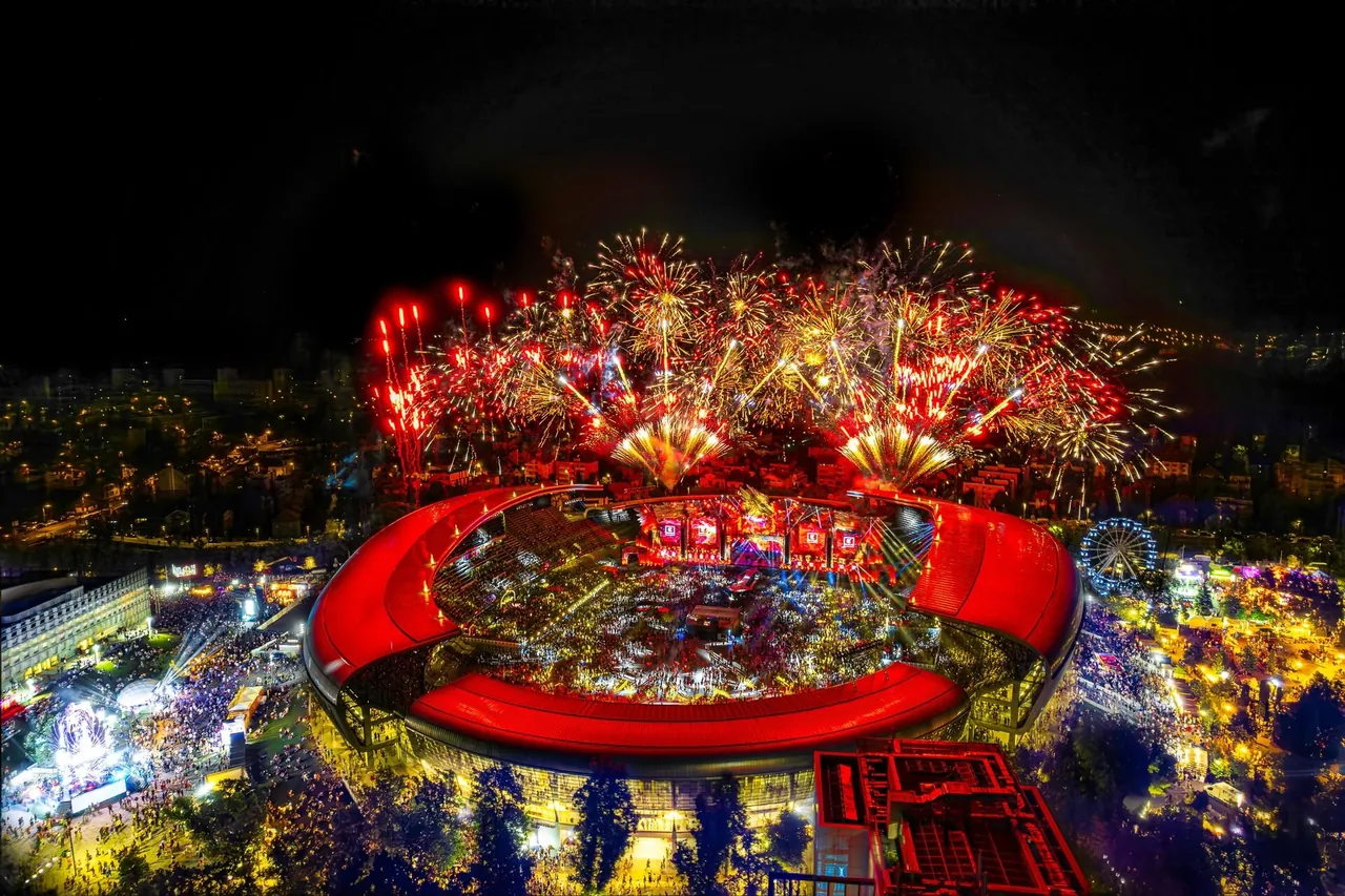 Vue aérienne de la Cluj Arena pendant un feu d'artifice, nuit du 5 au 6 août 2023 ; la grande roue et les scènes du parc central s'étendent autour du stade.