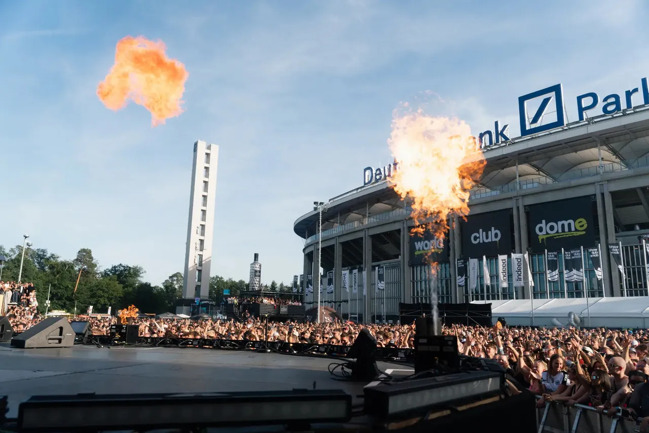Flame jets on a forecourt stage in front of the Deutsche Bank Park facade dressed in festival colours.