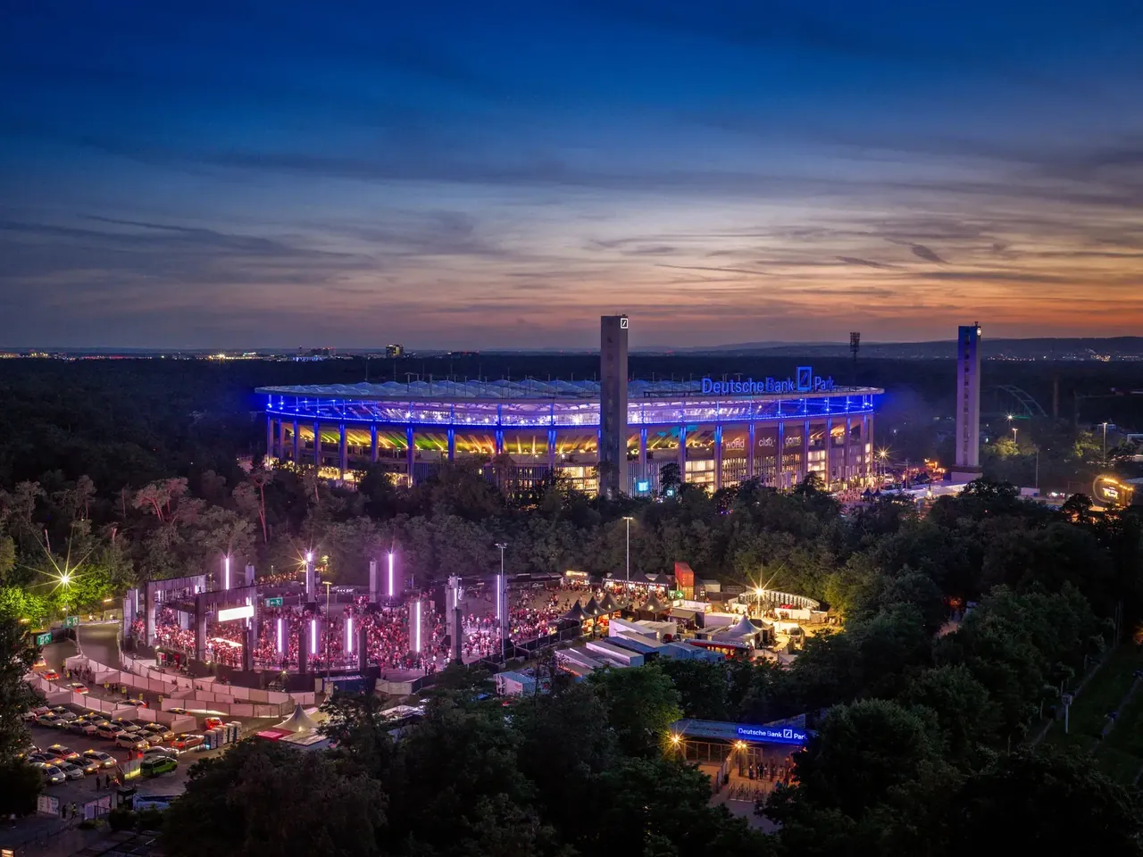 Deutsche Bank Park lit up at dusk in the middle of Frankfurt's city forest, with an illuminated outdoor stage in the foreground.