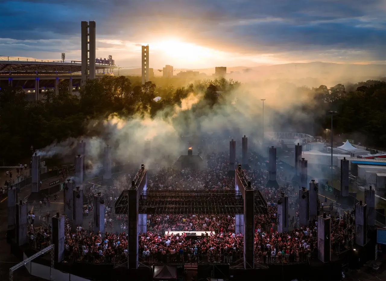 Elevated view of the UNREAL stage and its concrete monoliths at sunset, with the stadium and its two towers behind.