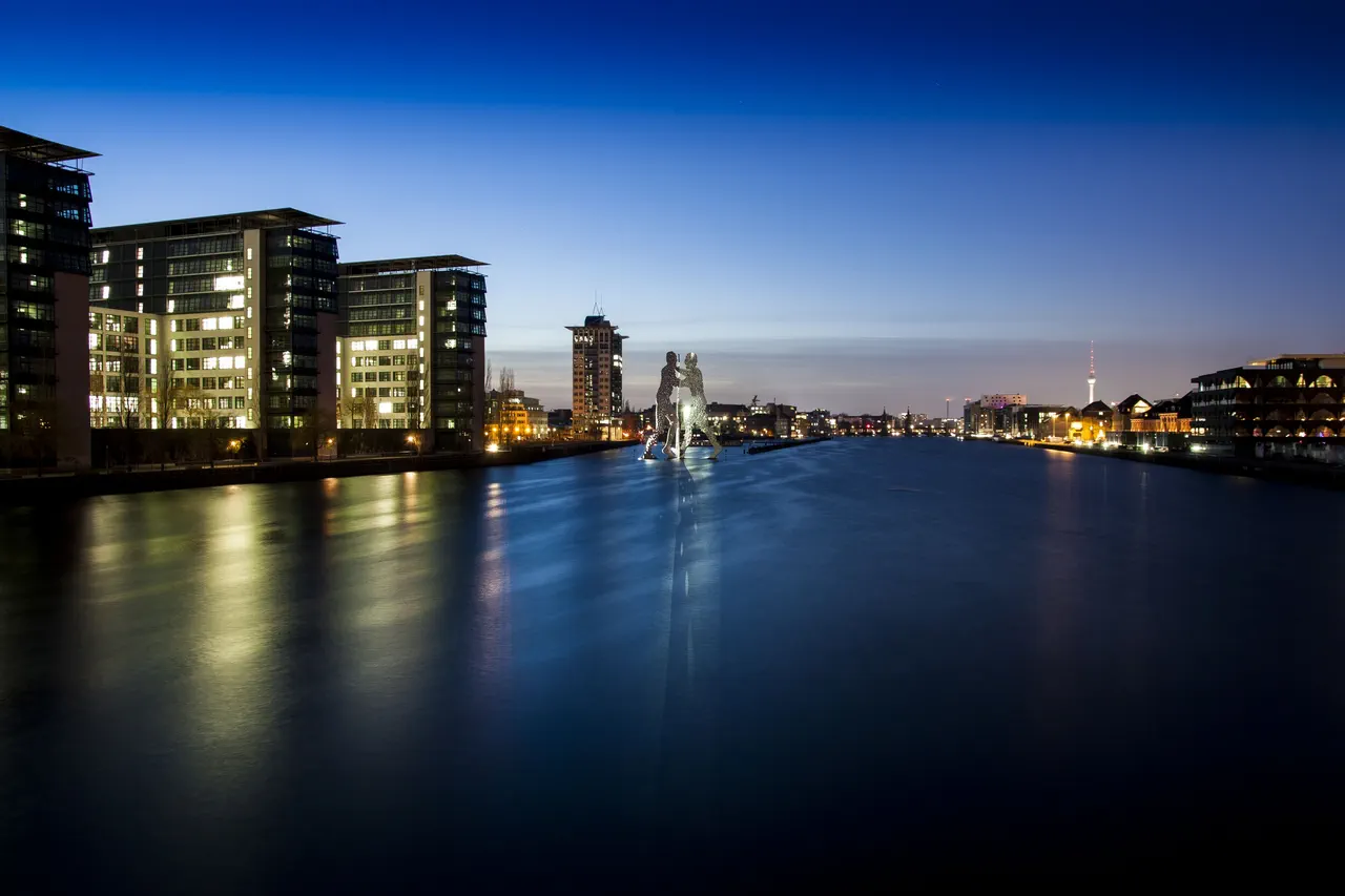 La Spree à Berlin à l’heure bleue, avec la sculpture Molecule Man au milieu de l’eau, des immeubles éclairés sur la rive et la tour de télévision au loin