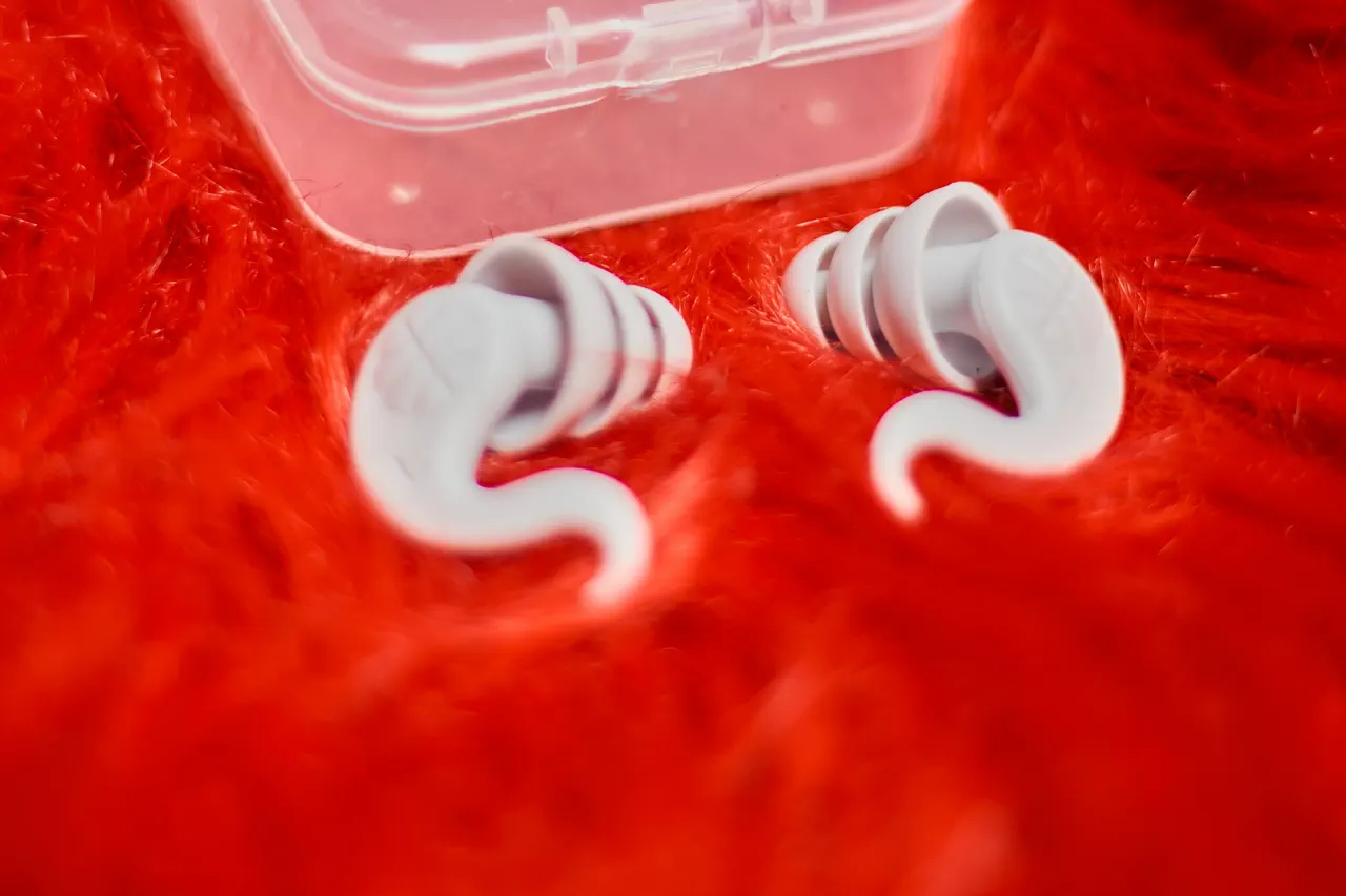 Paire de bouchons d’oreilles réutilisables en silicone gris clair posés sur une fourrure rouge, devant leur boîte transparente