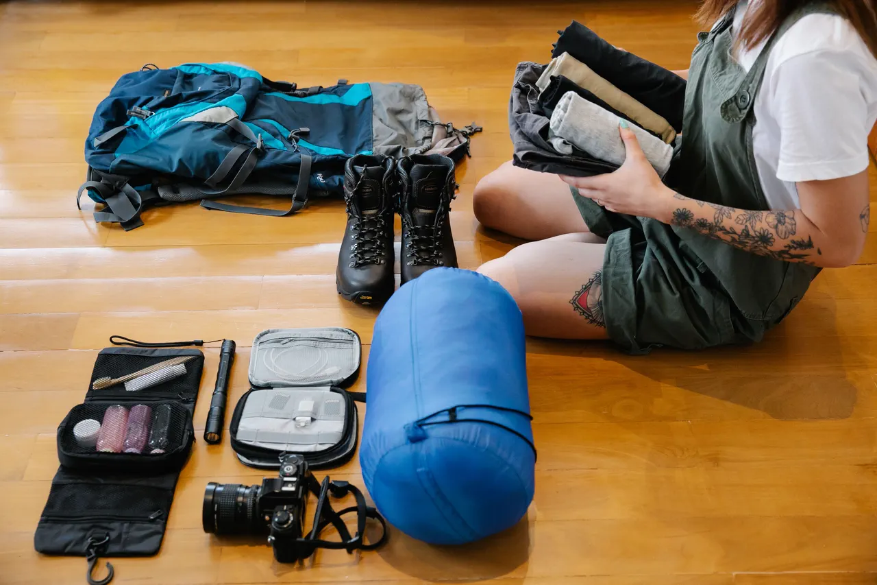 Affaires de voyage étalées sur un parquet : sac à dos, chaussures de randonnée, sac de couchage, trousse de toilette, lampe torche et appareil photo, à côté d’une personne qui plie des vêtements