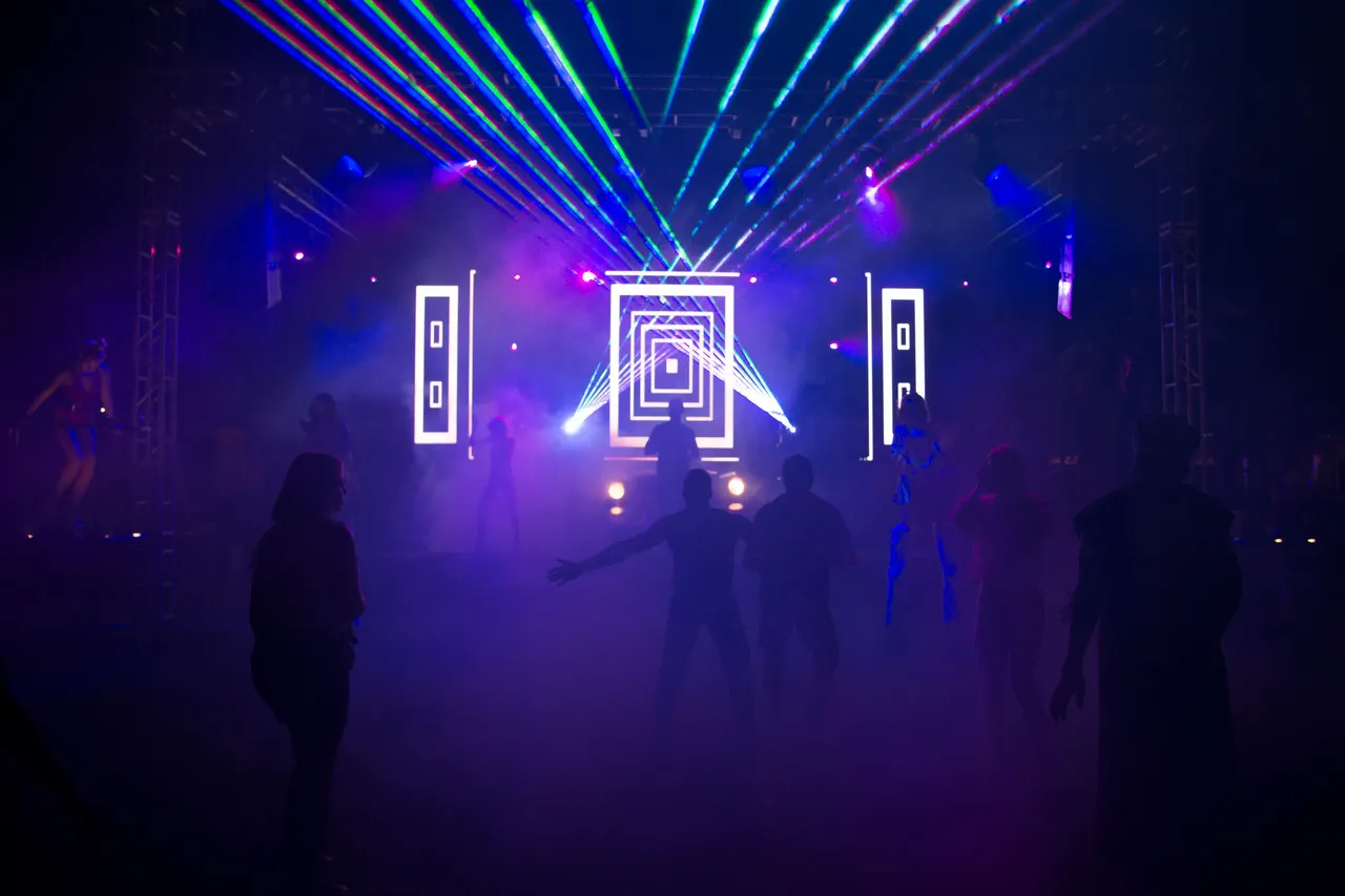 Silhouettes de danseurs dans la fumée, sous des faisceaux laser verts, bleus et roses, devant une scène à motifs lumineux