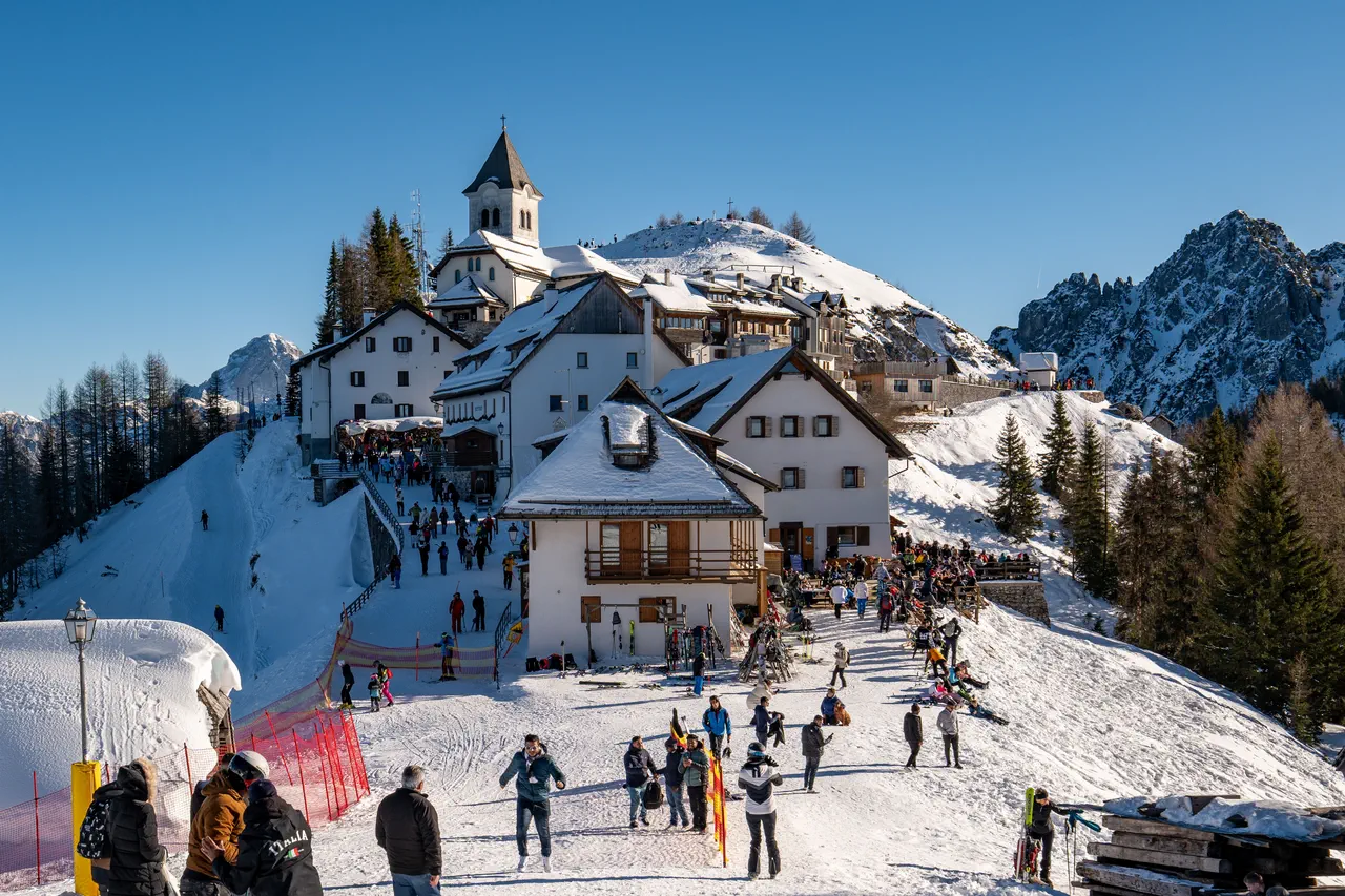 Village de montagne enneigé au sommet d’une piste, avec chalets, clocher et terrasses animées, skieurs et promeneurs sous un ciel bleu