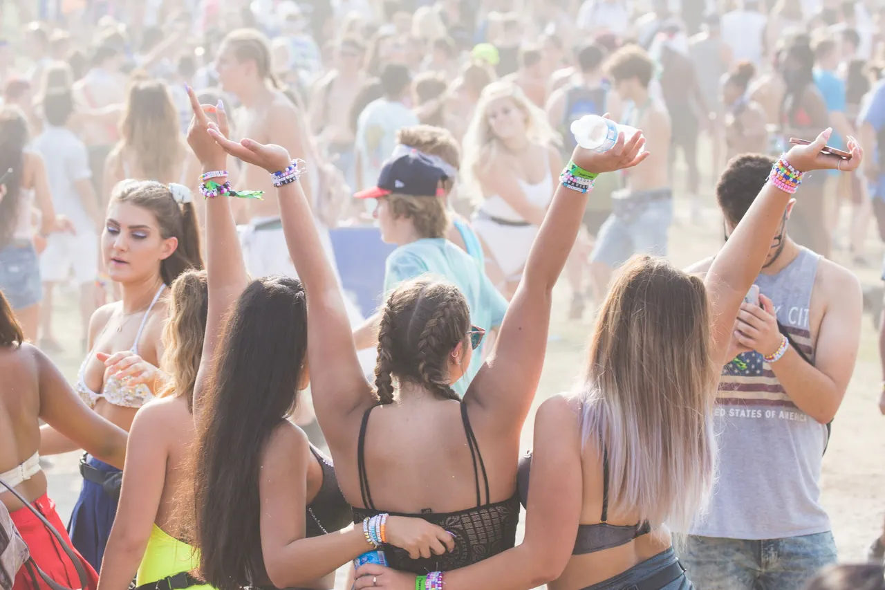 Festivaliers de dos, bras levés, en hauts courts et bracelets colorés, au milieu d’une foule en plein air par une journée ensoleillée