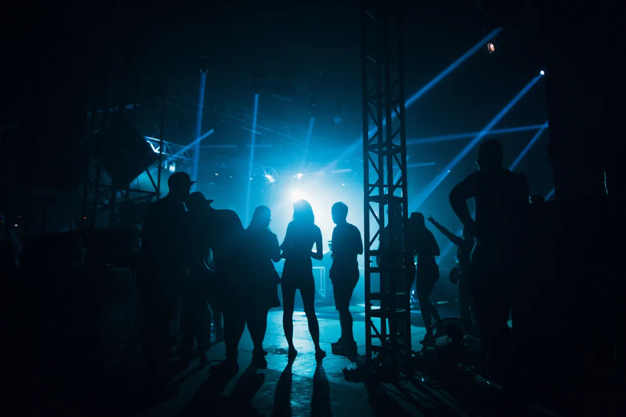 Groupe de personnes en silhouette, vêtues de sombre, dans une salle obscure traversée de faisceaux de lumière bleue